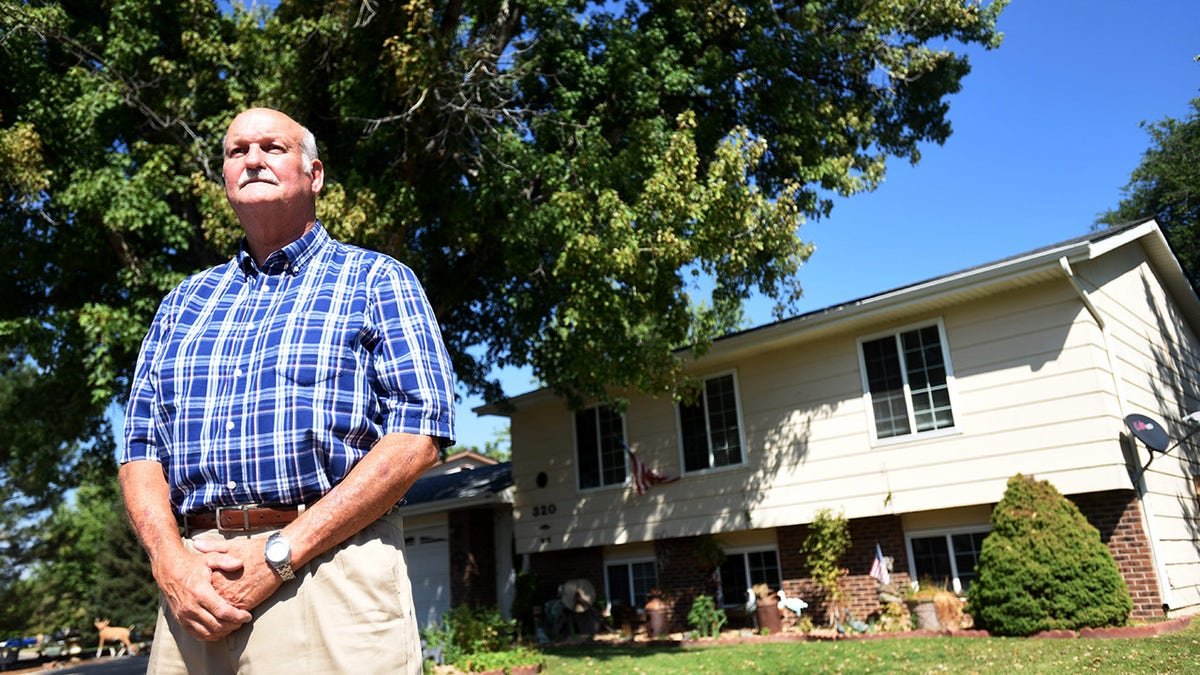 Russell Ross wearing a blue plaid shirt and khaki pants standing in front of a house.